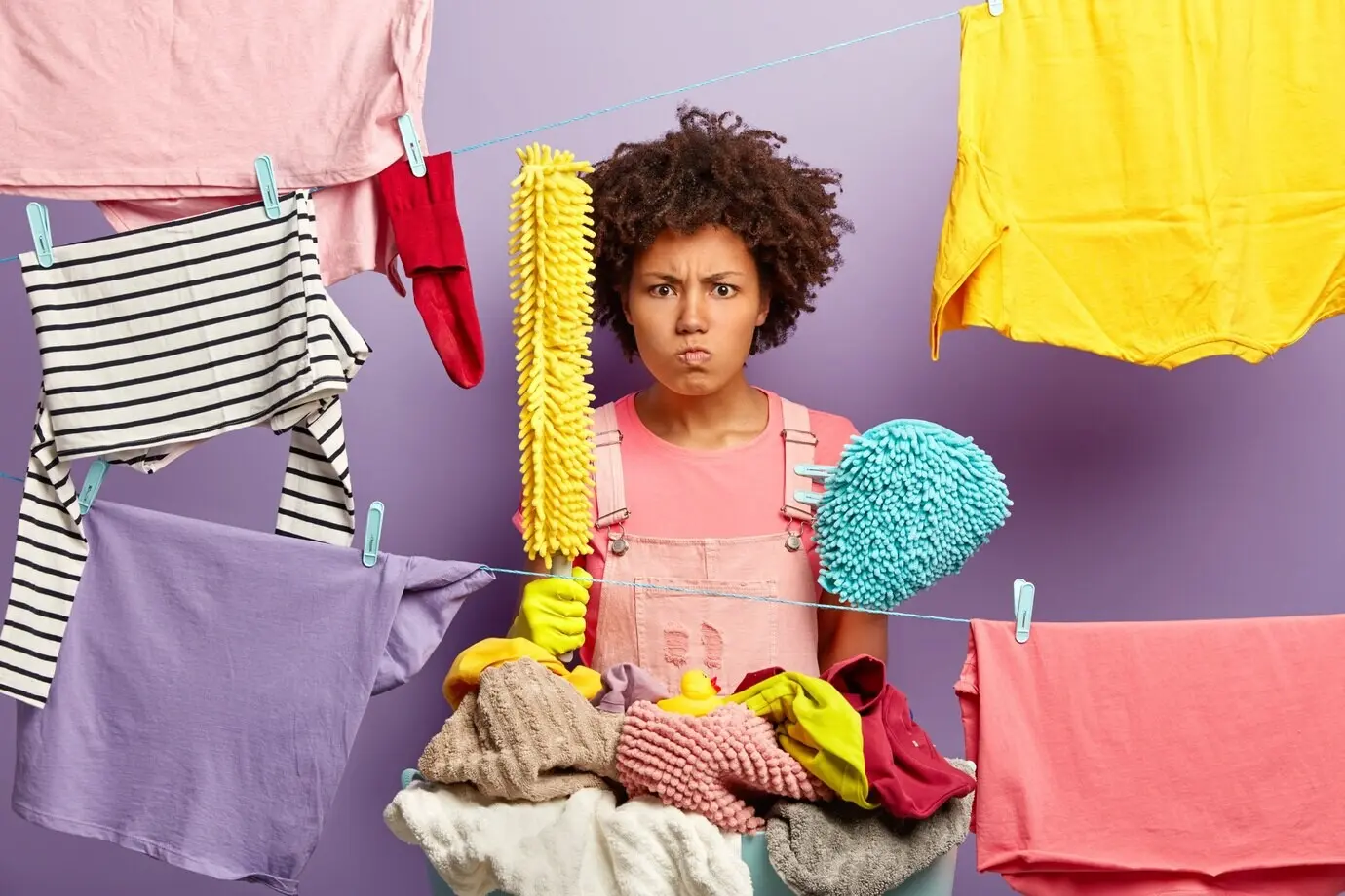 Раздраженная, недовольная женщина с прической афро держит принадлежности для стирки и стоит у бельевых верёвок с развешанной для сушки чистой одеждой; занята домашними делами и злится на ежедневные домашние обязанности. Концепция домашнего хозяйства.